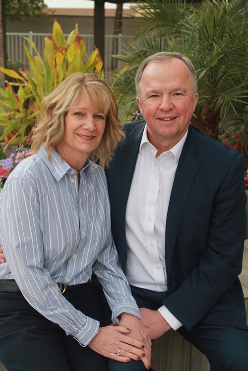 Skip and Traci Miller sitting in front of tropical plants smiling and holding hands